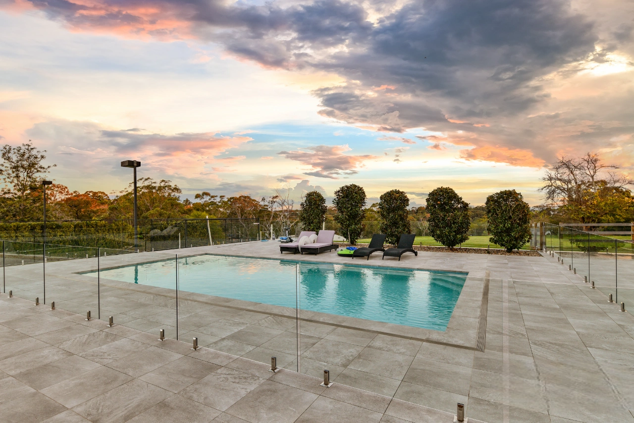 Luxury inground swimming pool with glass fence, large format tiled patio, and sun loungers set against a dramatic sunset sky.