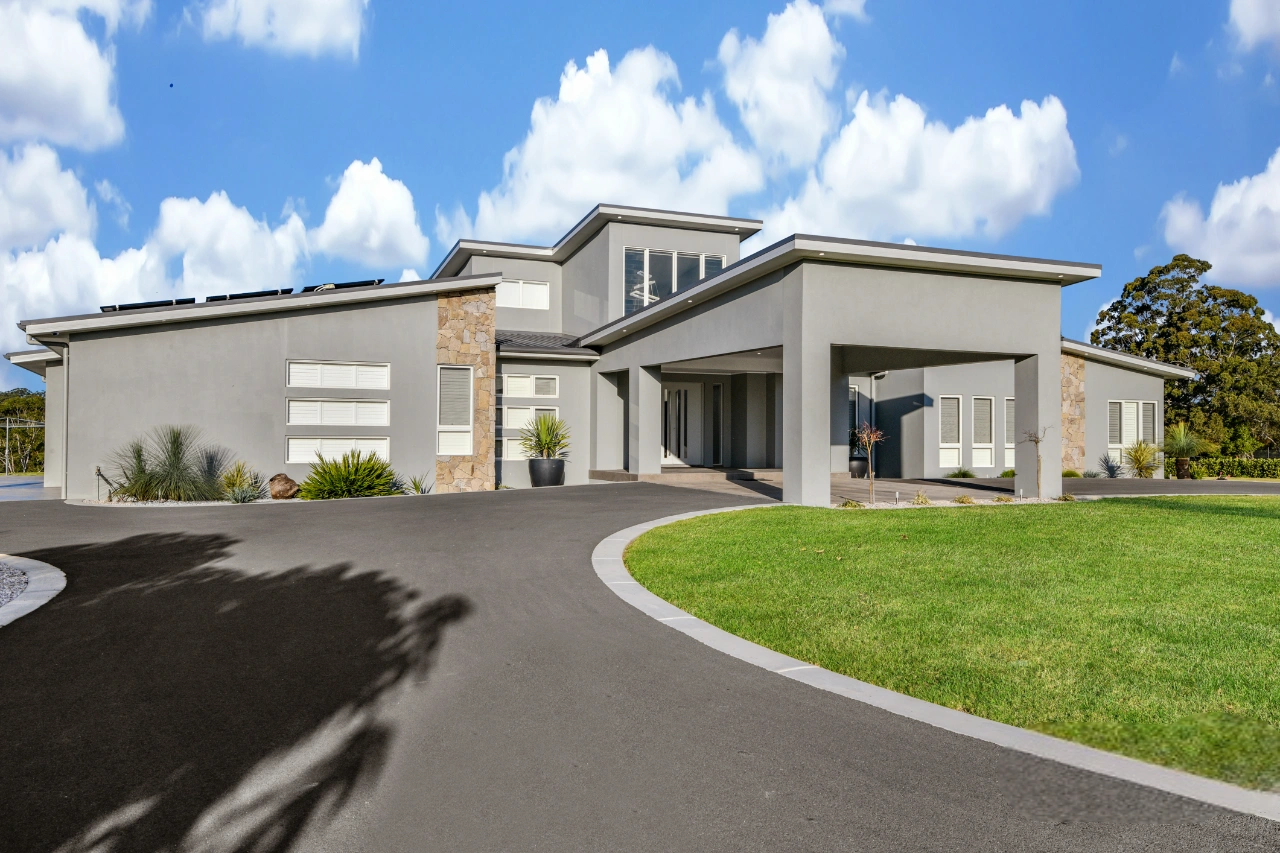 Large modern luxury home with gray stucco and stone veneer accents, featuring a porte-cochère and a long curved driveway on a sunny day.