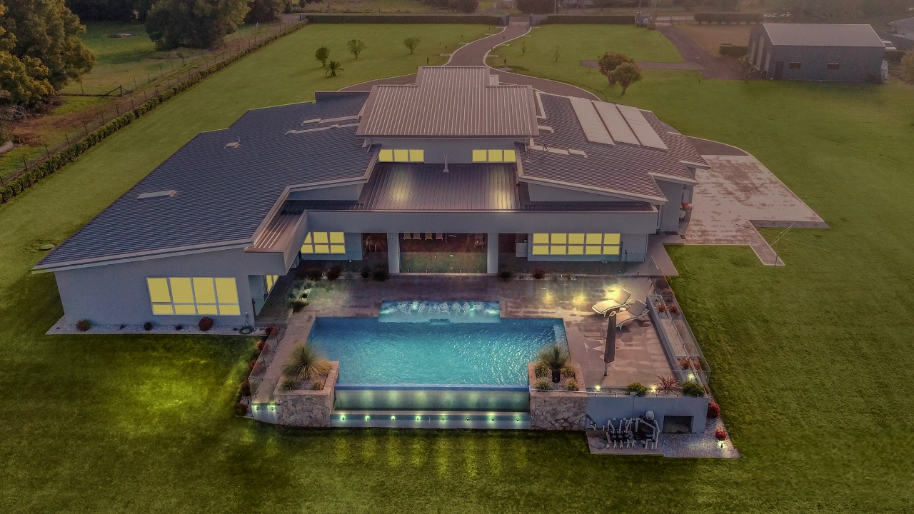 Aerial view of modern luxury estate with infinity pool, extensive lawn, and large contemporary house exterior lighting at dusk